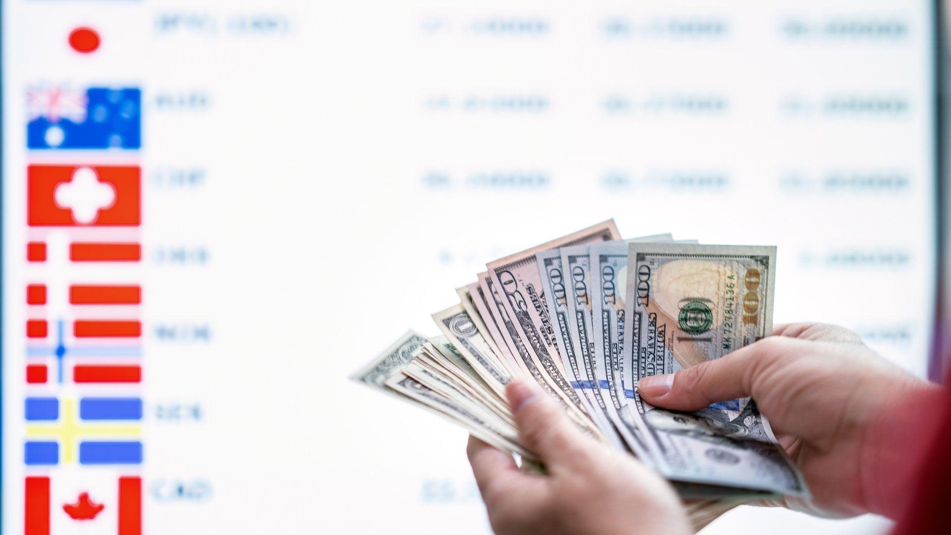 A hand holding US dollar banknotes in front of a currency rate board